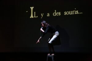 une comédienne sur un tabouret qui pointe le sol avec un écran derrière elle, où est écrit " Il y a des souris !"